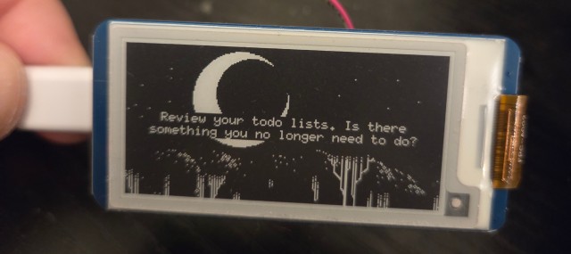 A small e-paper display, showing the words "Review your todo lists. Is there something you no longer need to do?". The text is superimposed over a 1-bit image of the top of a forest, at night, with a large crescent moon.